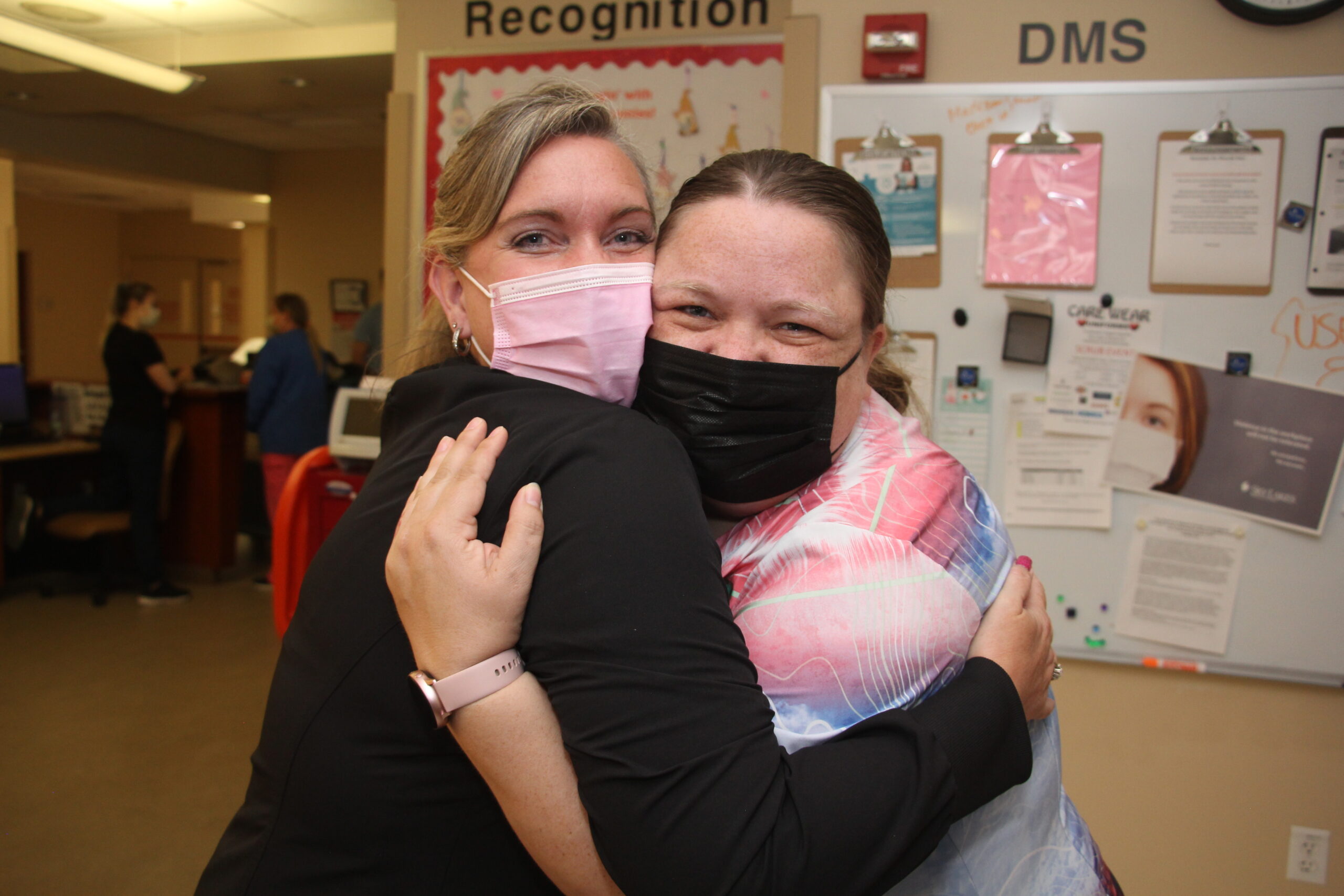 Two nurses in surgical masks smiling and hugging under in front of an information board that says "Recognition" and "DMS"